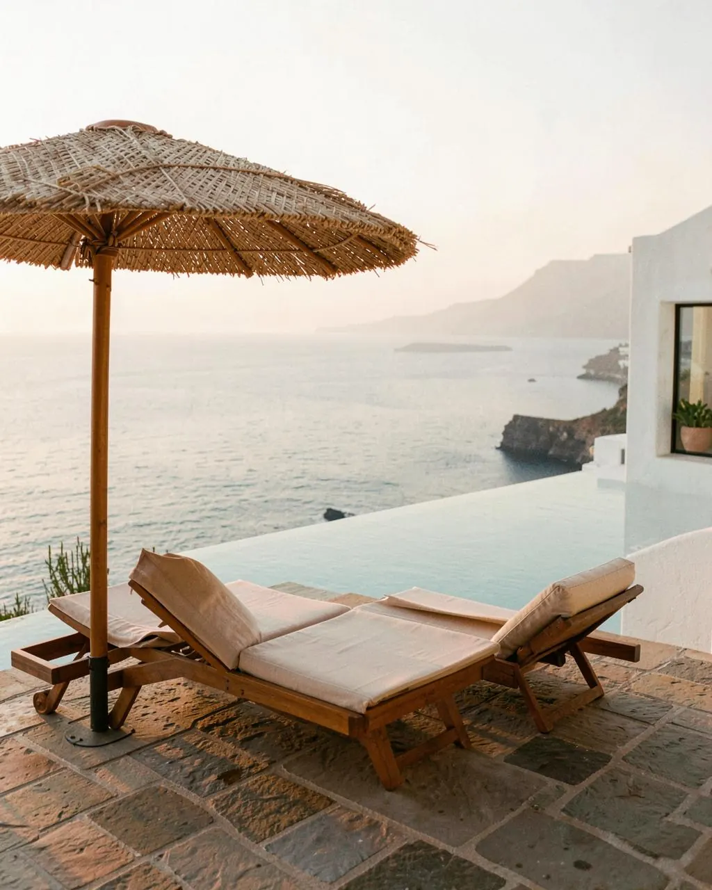 Two linen sun loungers under a raffia parasol beside an infinity pool overlooking a hazy Aegean sea at golden hour