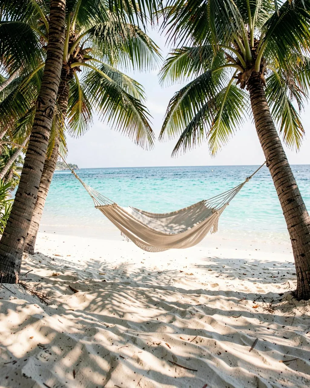A linen hammock strung between two palms on a white-sand cove above clear turquoise water