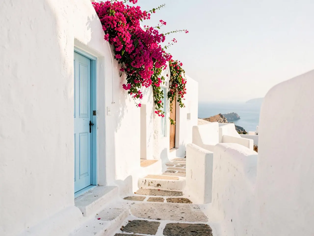 A whitewashed island lane with bougainvillea and a pale blue door, the sea glimpsed beyond